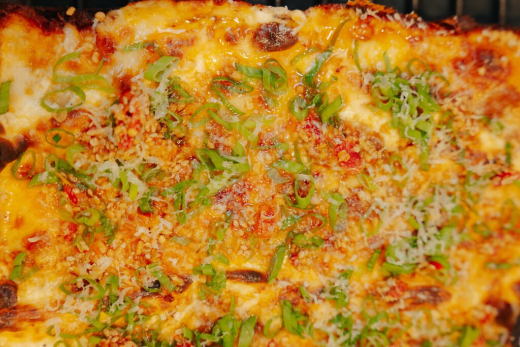 Close-up of a bubbly baked cheese casserole topped with melted cheese, red pepper flakes, and fresh sliced green onions for foodservice.
