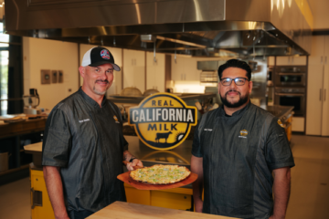 Two food service professionals holding a prepared dish in a commercial kitchen with Real California Milk branding visible.