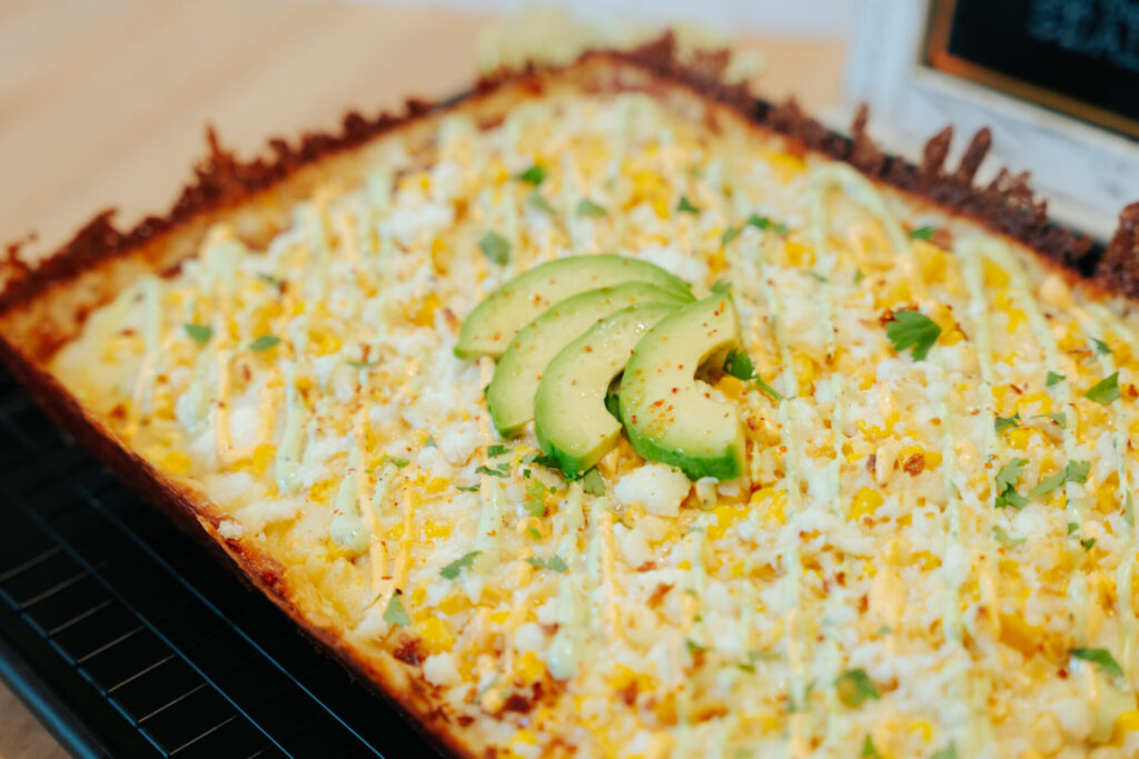Golden baked corn and cheese casserole topped with fresh avocado slices, cilantro, and spices in a rectangular dish.