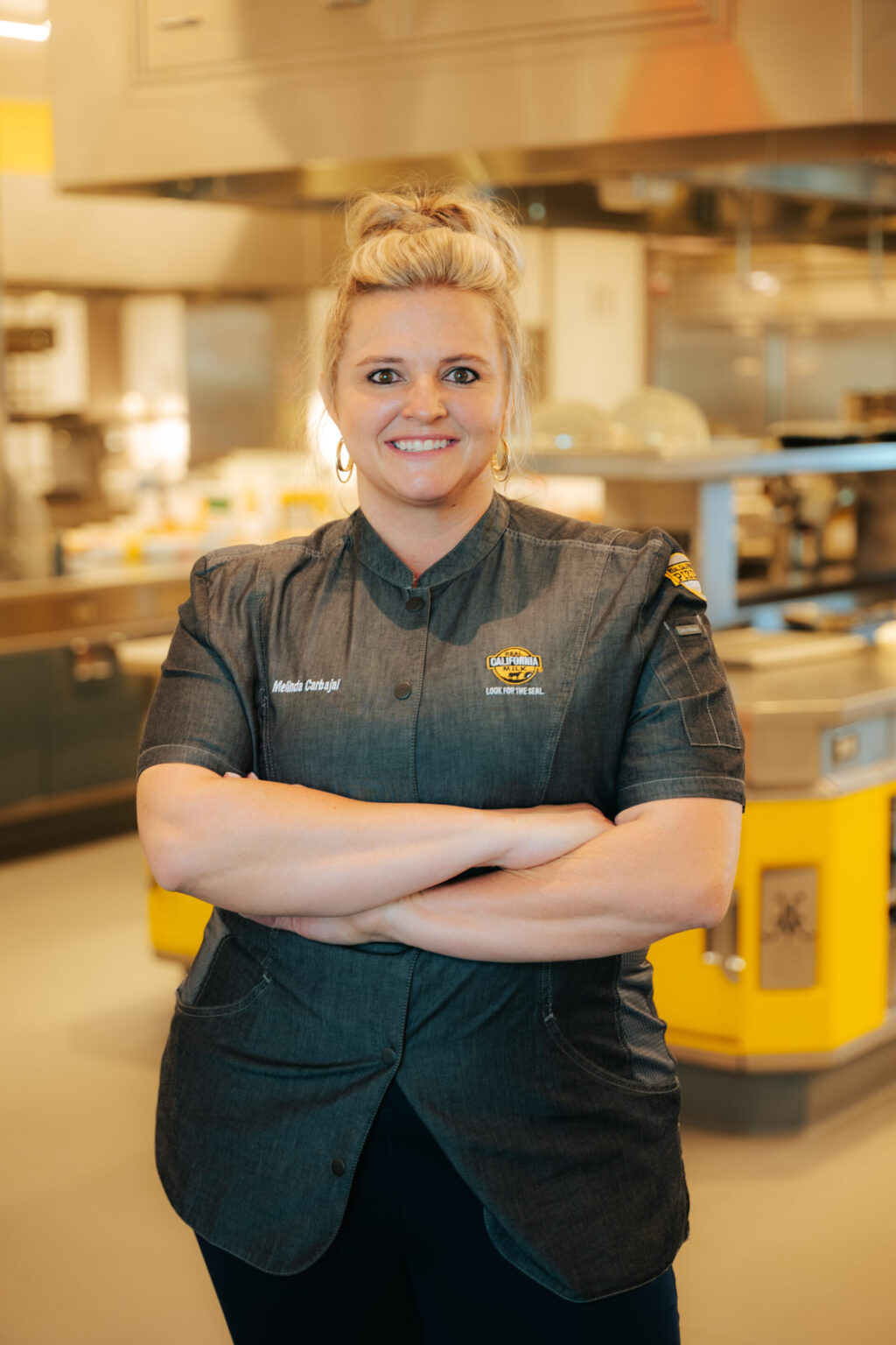 Female food service professional in denim uniform standing confidently in commercial kitchen facility with industrial equipment.
