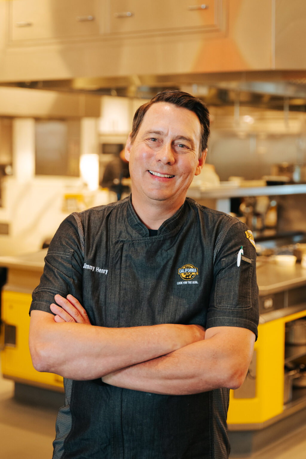 Portrait of a smiling food service worker in professional attire standing in a modern commercial kitchen facility.