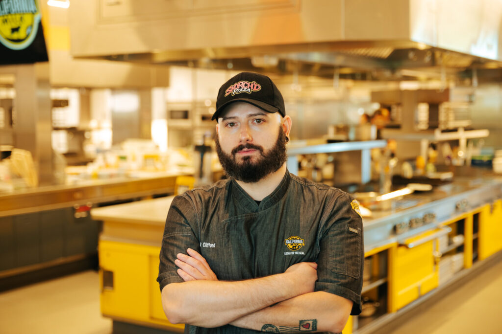 Food service worker in branded uniform standing in commercial warehouse with yellow safety equipment and processing facilities in background.