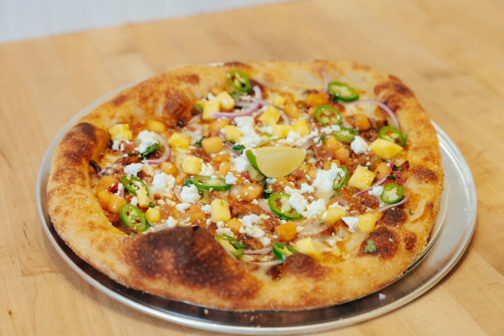 Artisan pizza topped with roasted corn, crumbled queso fresco, jalapeños, red onion, and lime wedge on wood surface.