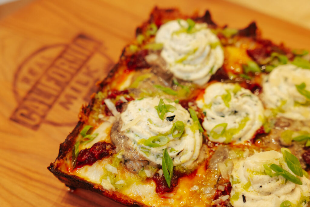Artisan pizza slice topped with melted cheese, cream cheese dollops, fresh green onions, and herbs on wooden surface.