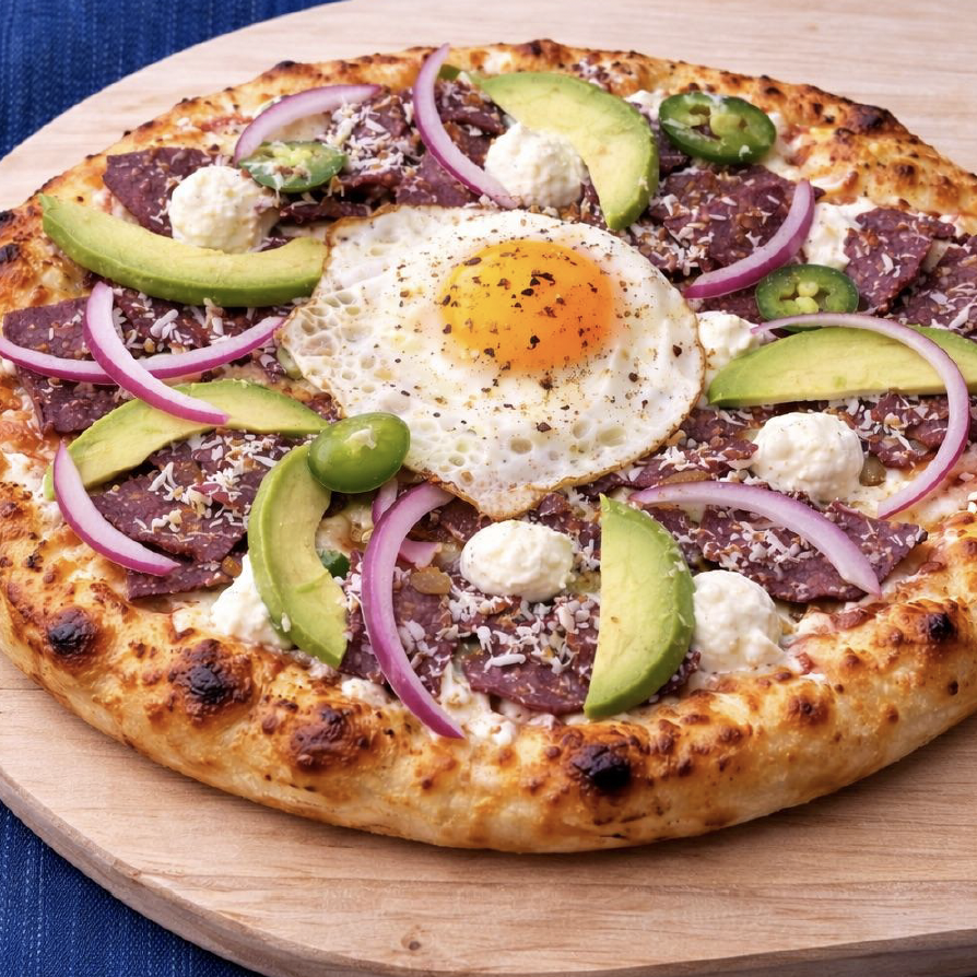 Artisan pizza topped with cured meat slices, fresh avocado, white cheese, red onion, and a sunny-side egg, ideal for upscale food service operations.