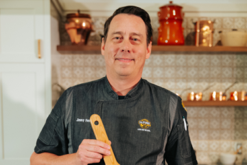 Professional chef in black chef coat holding wooden spoon in modern commercial kitchen with shelving and equipment.