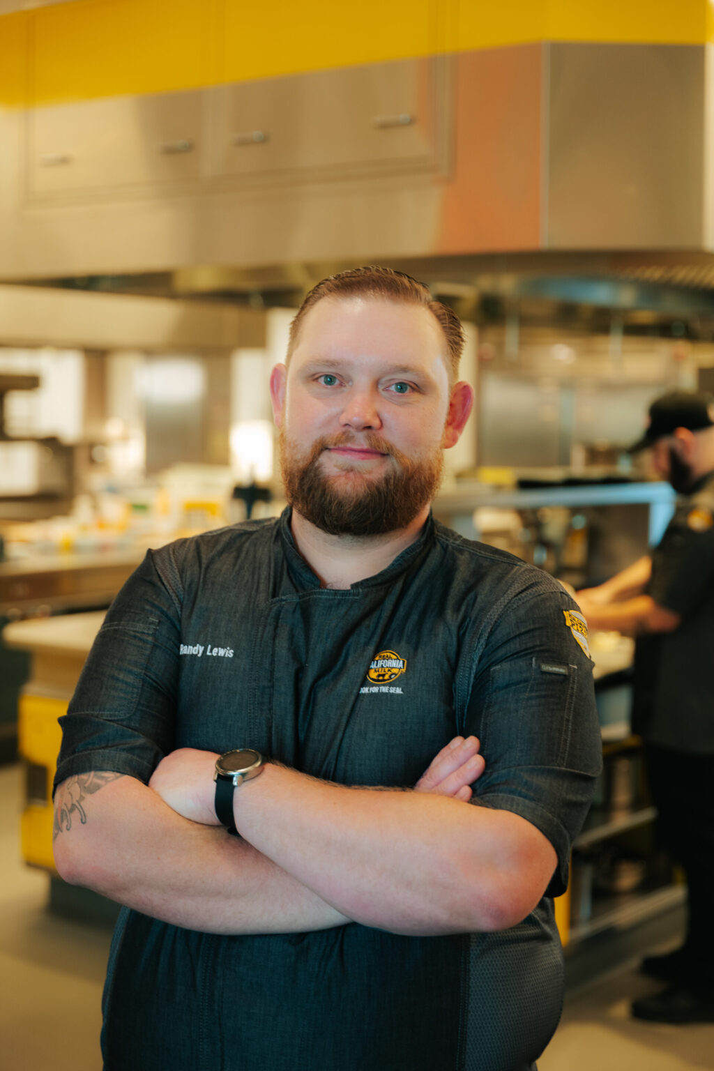 Experienced food service professional poses confidently in modern commercial kitchen facility with industrial equipment.