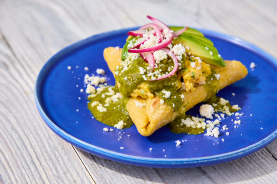 A blue plate with a Mexican empanada topped with green sauce, crumbled cheese, avocado slices, and pickled red onions, on a light wooden surface.