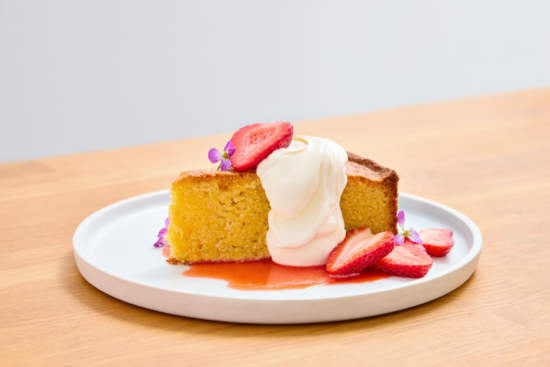 A slice of yellow cake topped with whipped cream, fresh strawberry slices, and small purple flowers sits on a white plate with strawberry sauce, on a light wooden table.