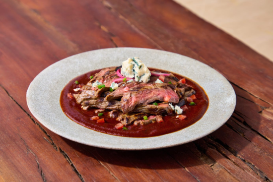 A plate of grilled steak slices topped with blue cheese and red onions, served over a rich red sauce with diced tomatoes and green herbs, on a rustic wooden table.