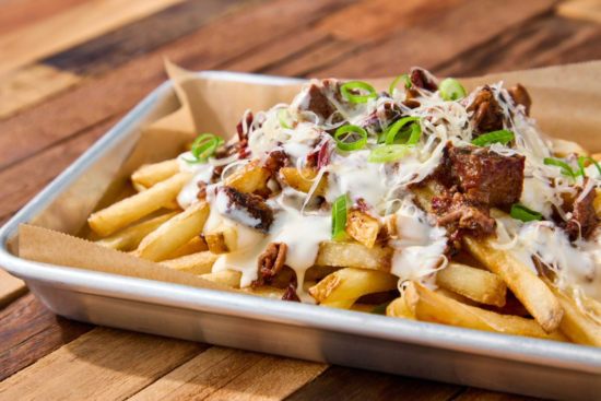 A metal tray lined with parchment paper holds French fries topped with shredded cheese, beef chunks, creamy sauce, chopped green onions, and pecans, set on a wooden table.