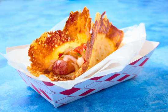 A hot dog topped with diced onions and tomatoes is served in a crispy, golden cheese shell, presented in a red-and-white paper tray against a blue background.