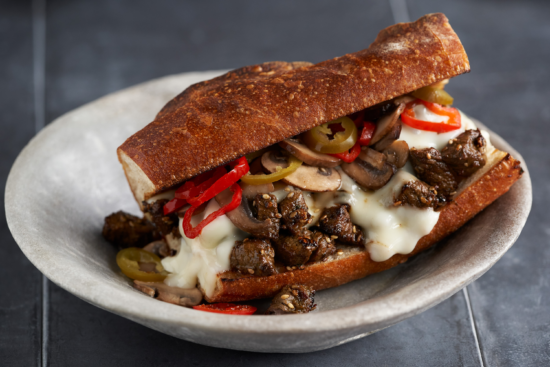 A toasted sandwich roll filled with melted cheese, sautéed beef, mushrooms, and sliced red and green peppers, served on a round ceramic plate.