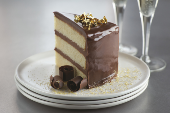 A slice of layered vanilla cake with glossy chocolate icing, topped with gold flakes and chocolate curls, sits on a white plate with gold sprinkles. Two glasses in the background suggest a celebratory setting.