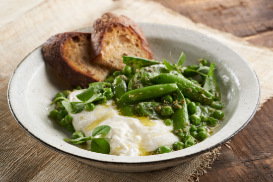 A rustic bowl with creamy burrata cheese, fresh green peas, snap peas, herbs, and a drizzle of olive oil, served with two slices of toasted bread on the side. The bowl rests on a wooden table.