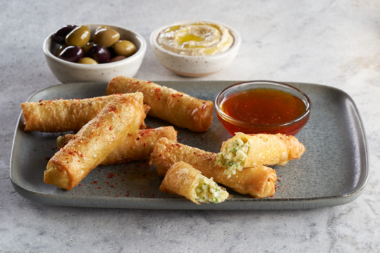 Six crispy rolls, some broken open to reveal a vegetable filling, on a gray plate with a bowl of red dipping sauce. In the background, bowls of mixed olives and hummus are visible.