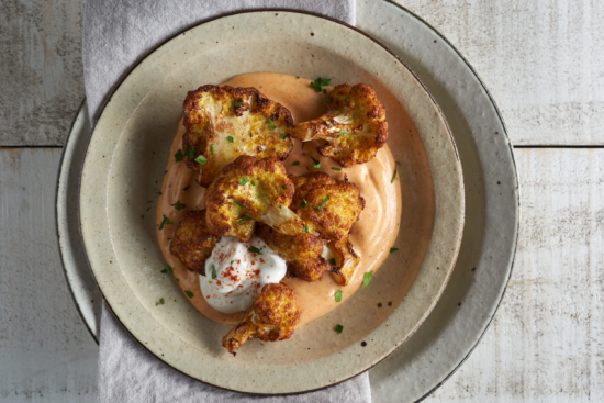 Roasted cauliflower florets served on a creamy orange sauce, topped with a dollop of white sauce and sprinkled with chopped herbs, on a rustic beige plate over a light napkin and wooden surface.