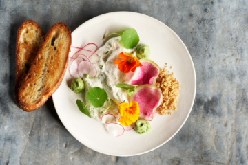 A white plate with toasted bread, herbed cheese spread, sliced radishes, green dollops, edible flowers, leafy greens, and a small pile of crumbled nuts on a gray surface.