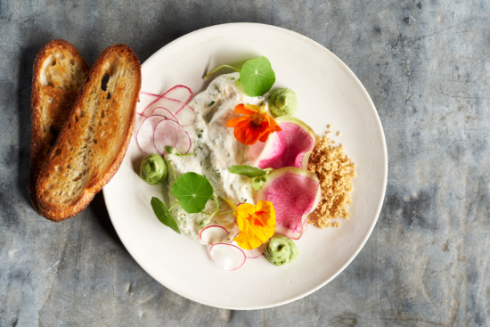 A white plate with toasted bread, herbed cheese spread, sliced radishes, green dollops, edible flowers, leafy greens, and a small pile of crumbled nuts on a gray surface.