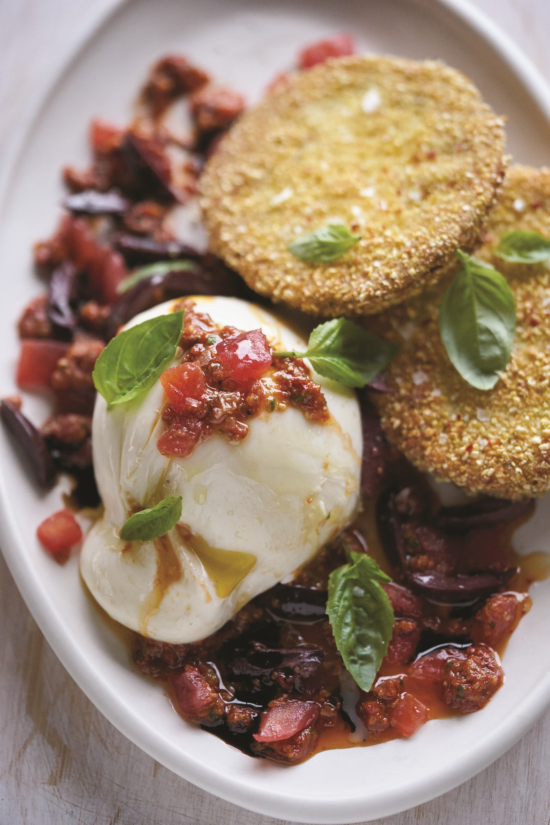 A plate featuring fried green tomato slices, a ball of burrata cheese topped with tomato relish, garnished with fresh basil leaves, all served on a white oval dish.