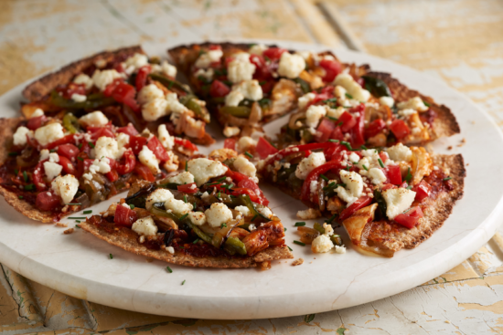 A round vegetable pizza topped with diced tomatoes, green bell peppers, onions, and crumbled cheese, sliced into six pieces and served on a white marble platter.