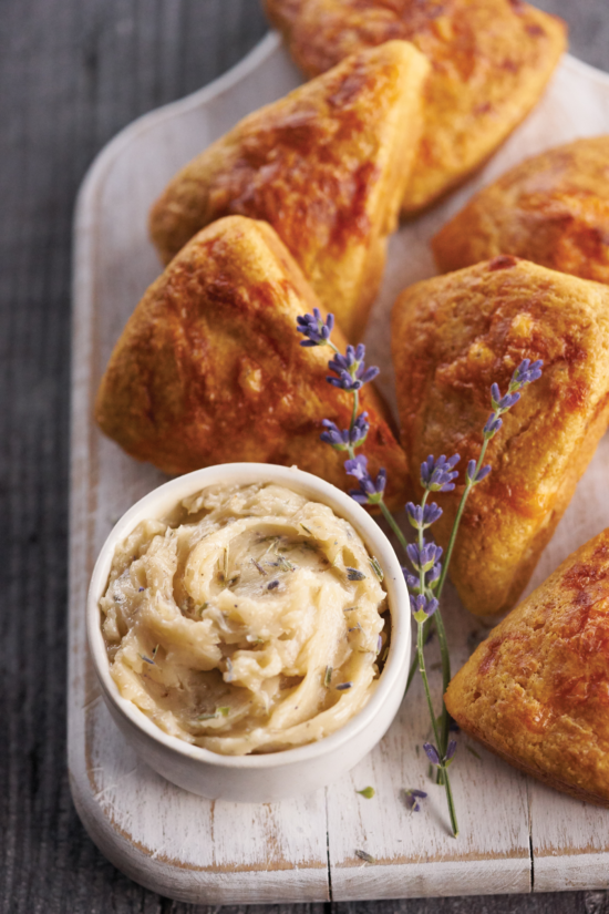 Golden scones on a white wooden board next to a small bowl of herb butter, garnished with sprigs of fresh lavender.