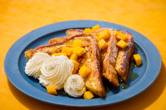 Three slices of French toast topped with diced mango and syrup, served with two swirls of whipped cream on a blue plate, set against a bright yellow background.