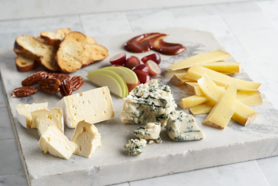 A marble board with assorted cheeses, sliced apples, grapes, pecans, sliced bread crisps, and fruit preserves arranged neatly.