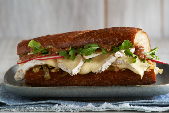 A close-up of a sandwich on a baguette with melted brie cheese, sliced apples, leafy greens, caramelized onions, and a spread, served on a plate over a blue cloth.
