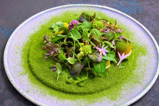A gourmet dish featuring assorted fresh greens, edible flowers, and vegetables, all artfully arranged on a round plate dusted with vibrant green powder, likely herb or matcha, creating a visually striking presentation.