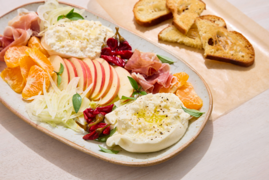 A platter with sliced apples, tangerine segments, fennel, prosciutto, spicy peppers, fresh burrata cheese drizzled with olive oil, and herbs, served with toasted bread slices on the side.
