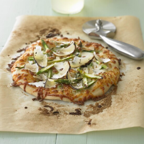 A small pizza topped with cheese, green peppers, mushrooms, and truffle shavings sits on parchment paper, with a pizza cutter placed nearby on a light surface.