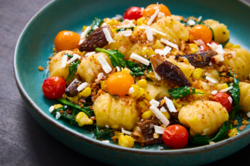 A blue plate filled with gnocchi, cherry tomatoes, spinach, corn, morel mushrooms, and crumbs, topped with grated cheese. The colorful ingredients are attractively arranged, creating a vibrant, appetizing dish.