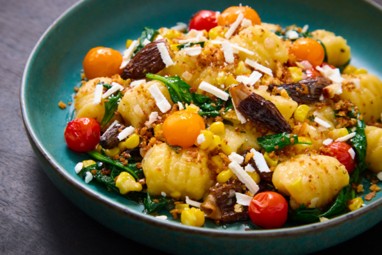 A blue plate filled with gnocchi, cherry tomatoes, spinach, corn, morel mushrooms, and crumbs, topped with grated cheese. The colorful ingredients are attractively arranged, creating a vibrant, appetizing dish.