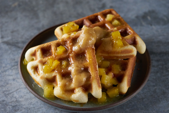 Two golden brown waffles topped with diced caramelized pineapple and syrup are served on a dark round plate, placed on a gray textured surface.