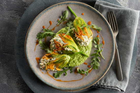 Three stuffed zucchini blossoms served on a light green sauce, garnished with arugula and a drizzle of red sauce, placed on a gray plate with a fork and napkin on the side.