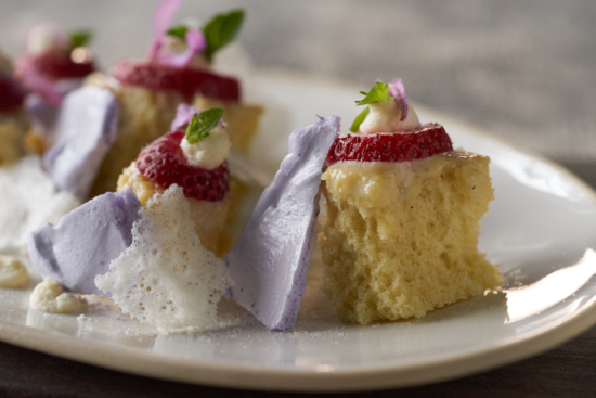 A close-up of a plated dessert featuring sponge cake pieces topped with whipped cream, strawberry slices, small green leaves, and purple meringue shards on a white plate.