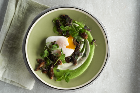 A gourmet dish featuring a soft-boiled egg, green pea purée, morel mushrooms, microgreens, and fresh herbs, artfully arranged on a light green plate set on a gray surface with a pale green napkin.