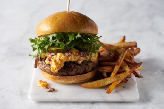 A cheeseburger topped with arugula, pickles, and a creamy spread, served on a bun with a side of golden French fries on a marble surface.