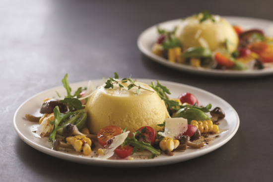 A plated dish featuring a yellow custard dome surrounded by arugula, cherry tomatoes, grilled corn, mushrooms, parmesan shavings, and microgreens. A second, similar plate is blurred in the background.