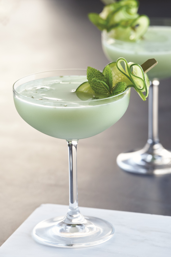 A pale green cocktail served in a coupe glass, garnished with cucumber slices and a sprig of mint. Another similar drink is blurred in the background. The scene is set on a light surface.