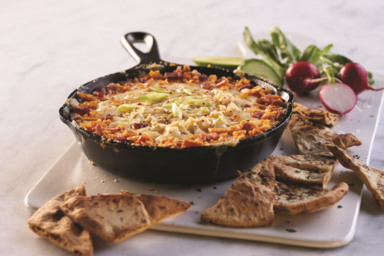 A skillet filled with baked cheesy dip topped with chopped green onions, surrounded by toasted pita bread slices, fresh radishes, lime wedges, and greens on a white serving board.