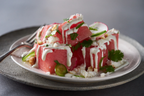 A plate of fresh watermelon cubes topped with white cheese, cream drizzle, sliced radishes, cilantro, and nuts, served with a fork on a ceramic plate.