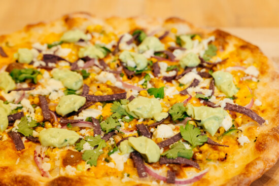 Vibrant pizza topped with guacamole, feta, corn, and colorful veggies, perfect for sharing.