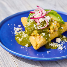 A blue plate with a Mexican empanada topped with green sauce, crumbled cheese, avocado slices, and pickled red onions, on a light wooden surface.