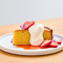 A slice of yellow cake topped with whipped cream, fresh strawberry slices, and small purple flowers sits on a white plate with strawberry sauce, on a light wooden table.
