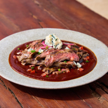 A plate of grilled steak slices topped with blue cheese and red onions, served over a rich red sauce with diced tomatoes and green herbs, on a rustic wooden table.