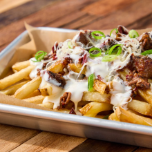 A metal tray lined with parchment paper holds French fries topped with shredded cheese, beef chunks, creamy sauce, chopped green onions, and pecans, set on a wooden table.