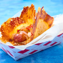 A hot dog topped with diced onions and tomatoes is served in a crispy, golden cheese shell, presented in a red-and-white paper tray against a blue background.