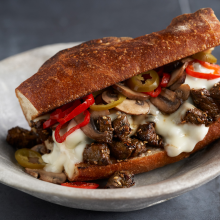 A toasted sandwich roll filled with melted cheese, sautéed beef, mushrooms, and sliced red and green peppers, served on a round ceramic plate.